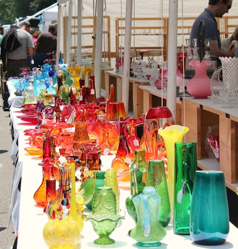 A collection of colorful glassware, including vases and decorative objects, is displayed on a long table at an outdoor market. The glass items come in a variety of vibrant colors such as red, orange, blue, green, and yellow, creating a visually striking display. Several people are browsing the market in the background.