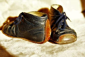 A pair of worn, vintage baby shoes with blue leather and brown soles. The shoes are laced with dark blue shoelaces, and the texture of the leather appears slightly cracked, suggesting age and use. The background is a soft, light-colored surface, enhancing the warm, nostalgic feeling of the shoes.