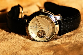 A luxurious wristwatch with a black leather strap and a metallic silver bezel. The watch face is adorned with Roman numerals and has an intricate design featuring a visible mechanical movement. It rests on a soft, textured, tan-colored fabric.