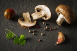 A composition of fresh ingredients including whole and sliced mushrooms, a cherry tomato, garlic clove, assorted peppercorns, and parsley leaves arranged on a dark slate surface.