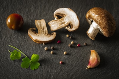 A composition of fresh ingredients including whole and sliced mushrooms, a cherry tomato, garlic clove, assorted peppercorns, and parsley leaves arranged on a dark slate surface.