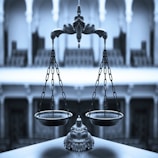 An ornate balance scale with intricate detailing is prominently positioned in the foreground against a blurred architectural backdrop of arches and columns, conveying a sense of elegance and precision.