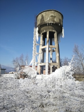 A weathered concrete water tower surrounded by a landscape covered in snow and icicles. Frost and ice hang from the tower's edges, and the ground is thick with white, frosted grass. Bare trees stand nearby, with a clear blue sky in the background.