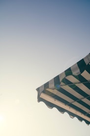 A striped awning is pictured against a clear sky, with the sun casting a soft glow and lens flare. The awning features alternating dark and light stripes, and the photograph conveys a sense of summer or outdoor dining.