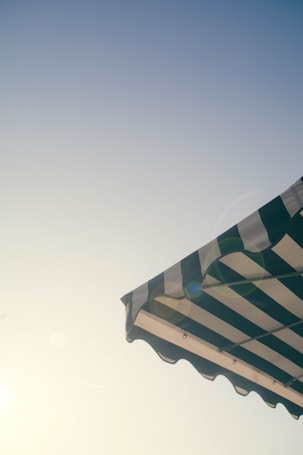 A striped awning is pictured against a clear sky, with the sun casting a soft glow and lens flare. The awning features alternating dark and light stripes, and the photograph conveys a sense of summer or outdoor dining.
