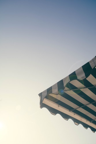 A striped awning is pictured against a clear sky, with the sun casting a soft glow and lens flare. The awning features alternating dark and light stripes, and the photograph conveys a sense of summer or outdoor dining.