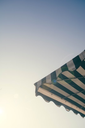 A striped awning is pictured against a clear sky, with the sun casting a soft glow and lens flare. The awning features alternating dark and light stripes, and the photograph conveys a sense of summer or outdoor dining.