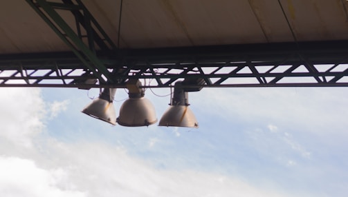 Three large industrial light fixtures are mounted on a metal framework under a roof. The sky in the background is partly cloudy, with light blues and whites.