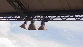 Three large industrial light fixtures are mounted on a metal framework under a roof. The sky in the background is partly cloudy, with light blues and whites.