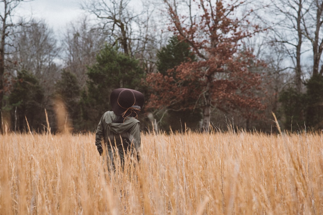 shallow focus photography of person wearing gray hooded jacket and black sun hat standing in brown grasses near green and brown leafed trees during daytime