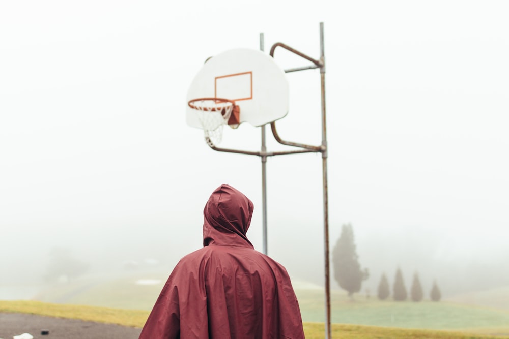 栗色のレインコートを着た人が、霧の深い昼間に白と灰色のバスケットボールのフープの下に立っています