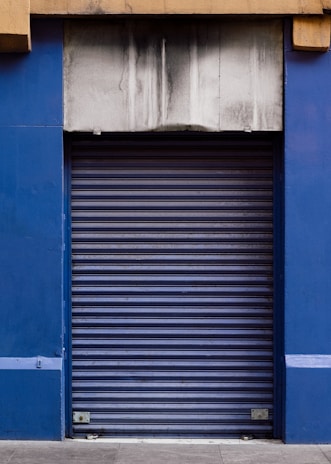 A closed blue rolling shutter door with a weathered and stained concrete facade above it. The structure is framed by blue walls on either side, and the ground is paved with concrete.