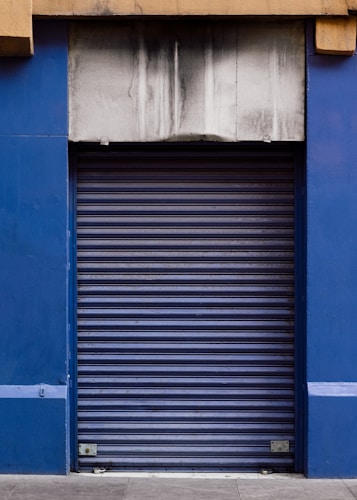 A closed blue rolling shutter door with a weathered and stained concrete facade above it. The structure is framed by blue walls on either side, and the ground is paved with concrete.