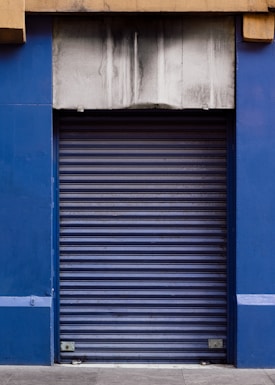 A closed blue rolling shutter door with a weathered and stained concrete facade above it. The structure is framed by blue walls on either side, and the ground is paved with concrete.