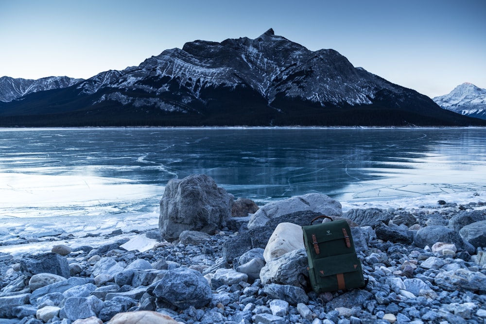 green and brown backpack leaning on stone near body of water