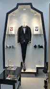 A display niche showcases a tuxedo jacket with a bow tie, centrally hung on a hanger. Surrounding the suit are glass shelves holding various items like perfumes and small boxes. The area is well-lit with overhead spotlights, highlighting the clothing and accessories. A glass table sits in front of the display, featuring magazines. The overall design is sleek and modern, with a black and white color scheme.