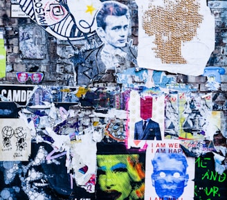 A vibrant urban collage on a brick wall featuring a mix of colorful posters, graffiti, and torn paper layers. Central elements include a monochrome portrait of a man with stylized hair, various abstract patterns, and vibrant, surreal imagery. Some posters have bold colors like neon green and pink, with text such as 'I AM WELL I AM HAPPY'.