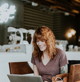 woman sitting on brown wooden chair while using silver laptop computer in room