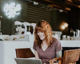 woman sitting on brown wooden chair while using silver laptop computer in room