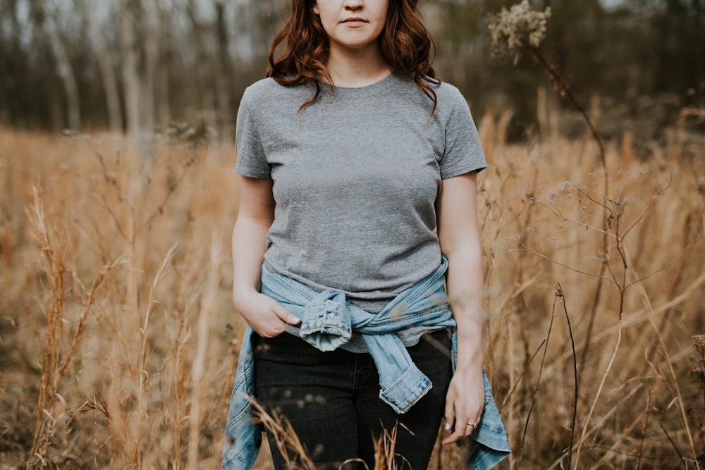 woman wearing gray crew-neck t-shirt with jacket wrap around her waist standing on brown grass field during daytime