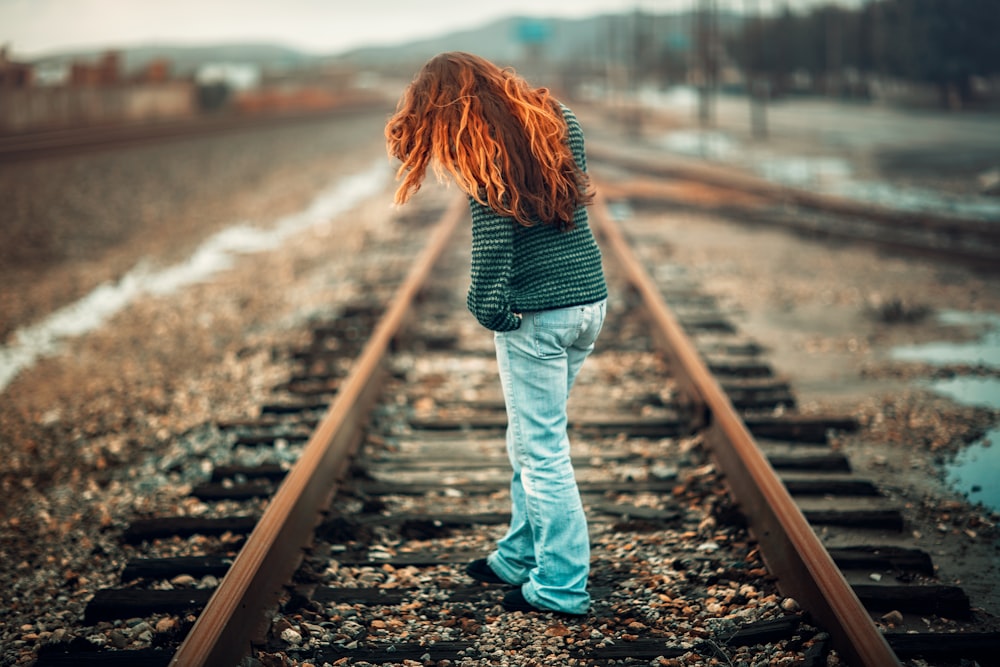 黒とグレーのストライプの長袖シャツと水色のデニムパンツを着たオレンジ色の髪の女性が、昼間、電車のレールの上に立っている