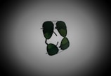A pair of aviator sunglasses with dark green lenses, prominently displayed against a gradient background. The sunglasses are positioned horizontally, with their reflection visible below.