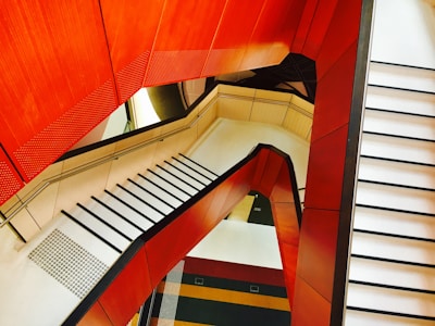 A modern and abstract architectural design featuring a staircase with vibrant red and orange accents. The angles and lines create a geometric pattern, providing a visually striking and dynamic perspective.