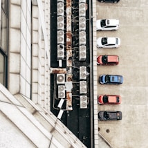 An aerial view of parked cars next to a building. The roof has a series of rusty air conditioning units aligned in a row. The cars are parked neatly in a parking lot with visible lines.