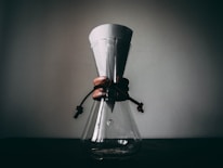 A glass coffee maker with a paper filter sits on a dark surface, featuring a wooden collar tied with leather straps. The background is a plain wall, creating a minimalistic and rustic atmosphere.