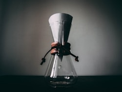 A glass coffee maker with a paper filter sits on a dark surface, featuring a wooden collar tied with leather straps. The background is a plain wall, creating a minimalistic and rustic atmosphere.