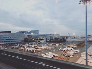An airport scene featuring multiple service vehicles parked in an organized manner on the tarmac. Buildings and roads are visible in the background under a cloudy sky, with bright lights illuminating parts of the area.