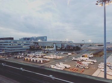 An airport scene featuring multiple service vehicles parked in an organized manner on the tarmac. Buildings and roads are visible in the background under a cloudy sky, with bright lights illuminating parts of the area.