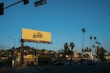 A street scene featuring a prominent yellow billboard with 'Los Feliz' written on it. The image includes various businesses such as 'Tang's Donuts & Ice Cream' and tall palm trees lining the street. Cars drive along the road under a clear blue sky with street signs reading 'Fountain' and 'Sunset'.