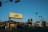 A street scene featuring a prominent yellow billboard with 'Los Feliz' written on it. The image includes various businesses such as 'Tang's Donuts & Ice Cream' and tall palm trees lining the street. Cars drive along the road under a clear blue sky with street signs reading 'Fountain' and 'Sunset'.