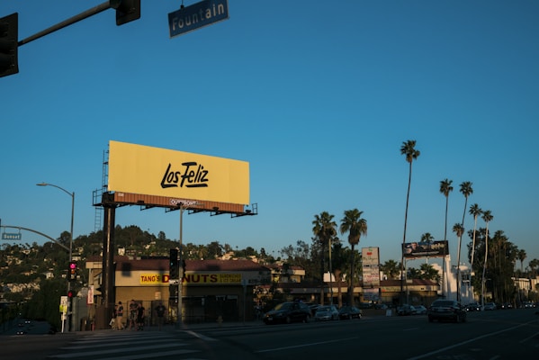 A street scene featuring a prominent yellow billboard with 'Los Feliz' written on it. The image includes various businesses such as 'Tang's Donuts & Ice Cream' and tall palm trees lining the street. Cars drive along the road under a clear blue sky with street signs reading 'Fountain' and 'Sunset'.