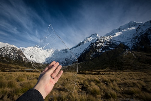 A hand holds a large, clear piece of ice, with snow-capped mountains and a grassy field in the background. The sky is partly cloudy, casting a serene light over the landscape.