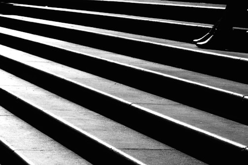 A high-contrast black and white photograph featuring a set of angular, sharply defined steps. The light and shadows create a dramatic pattern across the surface, with a solitary pair of feet visible in the upper right corner, suggesting motion.