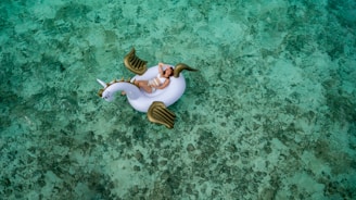 A person is lounging on an inflatable unicorn float in clear, shallow turquoise water. The water's surface displays a mix of light and darker green hues due to the sunlight and the sandy bottom. The unicorn float has white and gold elements, adding a whimsical touch to the serene setting.