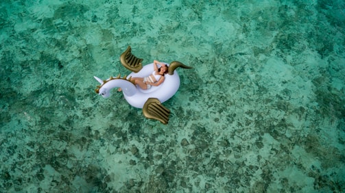 A person is lounging on an inflatable unicorn float in clear, shallow turquoise water. The water's surface displays a mix of light and darker green hues due to the sunlight and the sandy bottom. The unicorn float has white and gold elements, adding a whimsical touch to the serene setting.