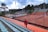 An empty outdoor tennis court surrounded by rows of colorful stadium seating, with a person in a red jacket climbing the orange seats. The court is enclosed by a fence, and there are several tall light poles and palm trees in the background under a cloudy sky.