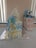 A diaper cake made of stacked rolled diapers decorated with blue and yellow ribbons, topped with a white cloth. Beside it, there is a gift bag with a green and pink floral pattern, containing a blue stuffed bear toy. The setup is placed on a table covered with a white cloth.