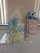 A diaper cake made of stacked rolled diapers decorated with blue and yellow ribbons, topped with a white cloth. Beside it, there is a gift bag with a green and pink floral pattern, containing a blue stuffed bear toy. The setup is placed on a table covered with a white cloth.