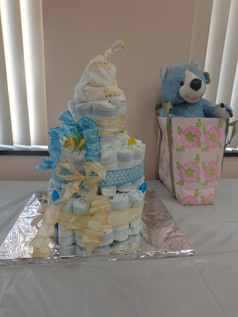 A diaper cake made of stacked rolled diapers decorated with blue and yellow ribbons, topped with a white cloth. Beside it, there is a gift bag with a green and pink floral pattern, containing a blue stuffed bear toy. The setup is placed on a table covered with a white cloth.