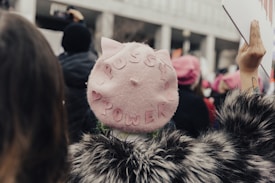 A person wearing a pink hat with the words 'Pussy Power' stitched on it, surrounded by a crowd of people. The person is wearing a faux fur coat, and there are signs held up in the background. The scene appears to be a gathering or protest with a focus on empowerment.