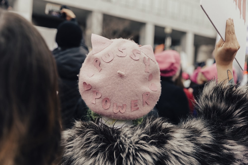 selective focus photography of person wearing pink fleece Pussy Power cap raising right hand