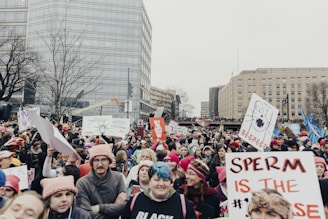 A large, diverse crowd of people marches through a city street, many holding various signs advocating for rights and social issues. The crowd is densely packed and spans a wide area, suggesting a protest or rally. There are modern buildings in the background, and some trees without leaves, indicating a winter setting. Many participants are wearing pink hats, and some signs display messages promoting respect and freedom.