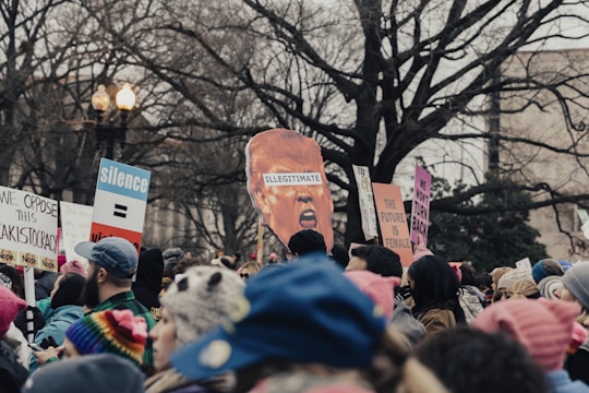 A large group of people are gathered in a public space, holding signs with various political messages and slogans. Some signs contain statements like 'The Future is Female' and 'We Oppose This Kakistocracy'. The crowd is diverse, with many people wearing colorful winter clothing. A prominent cutout of a head labeled 'Illegitimate' is also seen among the crowd. Bare trees and a lamp post frame the background.