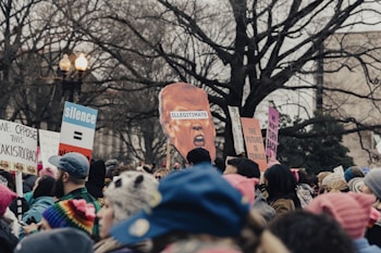 A large group of people are gathered in a public space, holding signs with various political messages and slogans. Some signs contain statements like 'The Future is Female' and 'We Oppose This Kakistocracy'. The crowd is diverse, with many people wearing colorful winter clothing. A prominent cutout of a head labeled 'Illegitimate' is also seen among the crowd. Bare trees and a lamp post frame the background.