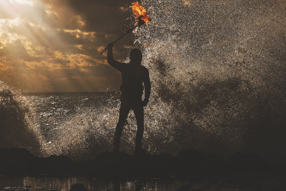 A man standing in the water during sunset at dusk, while holding a torch.