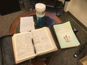 An open Bible and a book titled 'Becoming a Millionaire God's Way' are placed on a wooden table. There is a cup with the Starbucks logo, handwritten notes on a notecard, a pen resting on the Bible, and a black leather chair partially visible in the background.